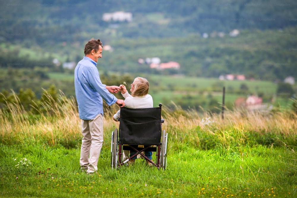 Hombre mayor con mujer sentada en silla de ruedas, tomados de la mano, oustide en la naturaleza verde del otoño, vista trasera