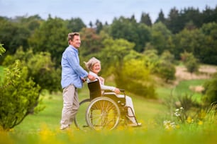 Homem sênior que empurra a mulher sentada na cadeira de rodas oustide na natureza verde do outono