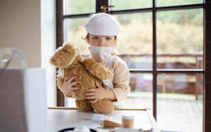 Niña pequeña con uniforme de médico en el interior de su casa, jugando con un oso de peluche.