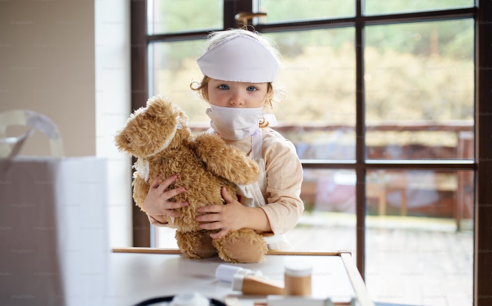Kleines Kleinkind Mädchen mit Arztuniform drinnen zu Hause, spielt mit Teddybär.