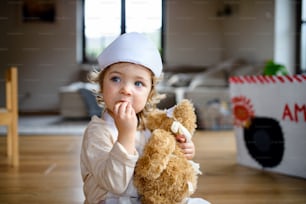 Menina pequena com uniforme médico dentro de casa, brincando com ursinho de pelúcia.