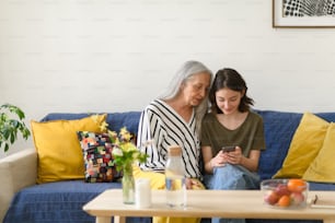 Una abuela mayor feliz con un abuelo adolescente sentado en el sofá y usando teléfonos inteligentes juntos en casa.