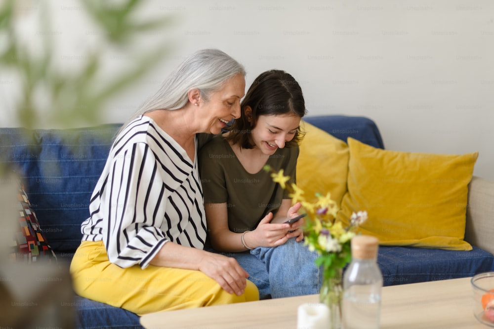 A happy senior grandmother with teenage granddaguhter sitting on sofa and using smartphone together at home.