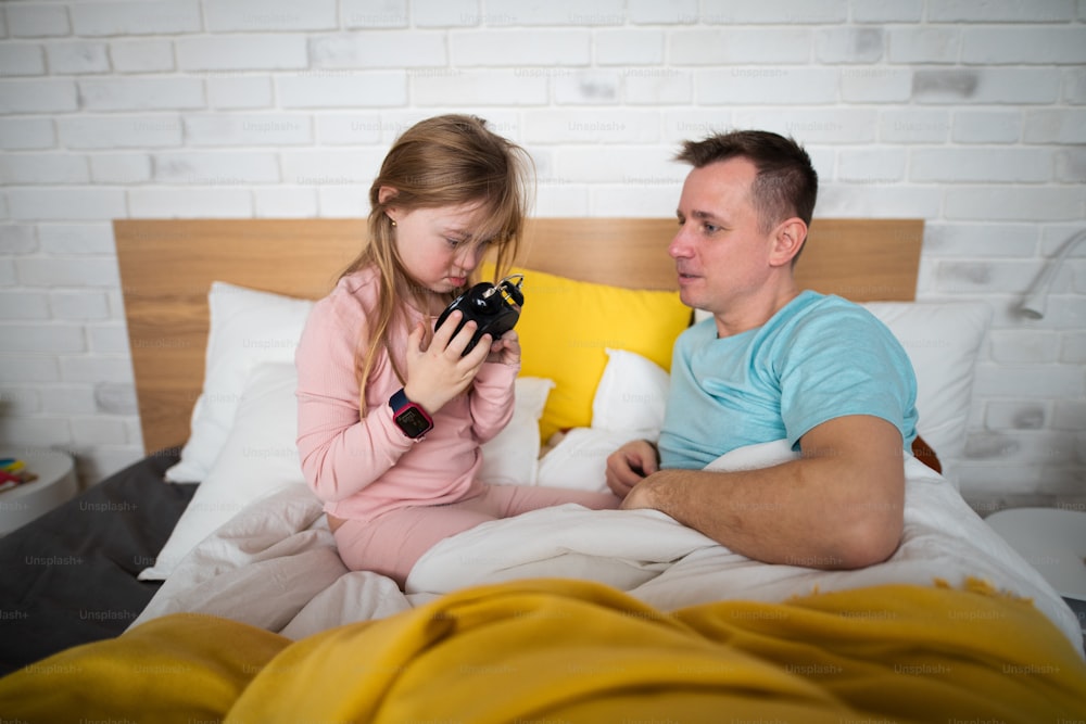 A father setting alarm clock with his little daughter with Down syndrome in bed at home.