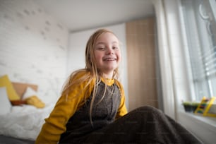 Ein glückliches kleines Mädchen mit Down-Syndrom, das zu Hause auf dem Bett sitzt.