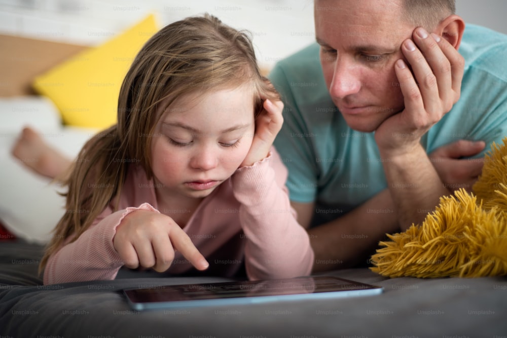 ダウン症の父と幼い娘がベッドに横たわり、家でタブレットを使っている。