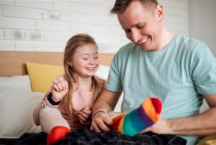 A father putting on different socks to his little daughter with Down syndrome when sitting on bed at home.