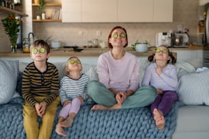 Three little children with a mother sitting on sofa and putting cucumber on faces at home.