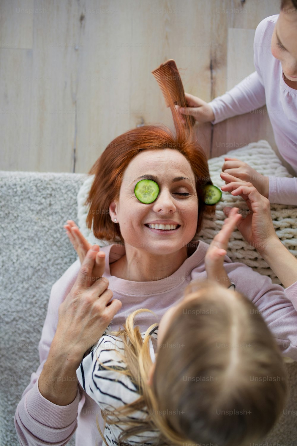 Two little girls putting a cucumber on their mother's face and combing her hair at home.