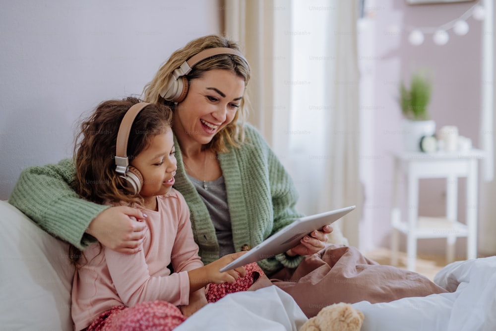 A happy mother with her little daughter sitting on bed and using tablet and headphones at home.