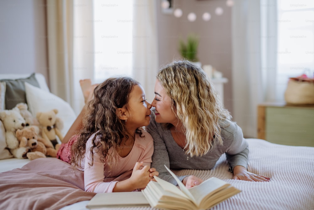 A happy mother with her little daughter lying on bed and tlooking at each other when reading book.