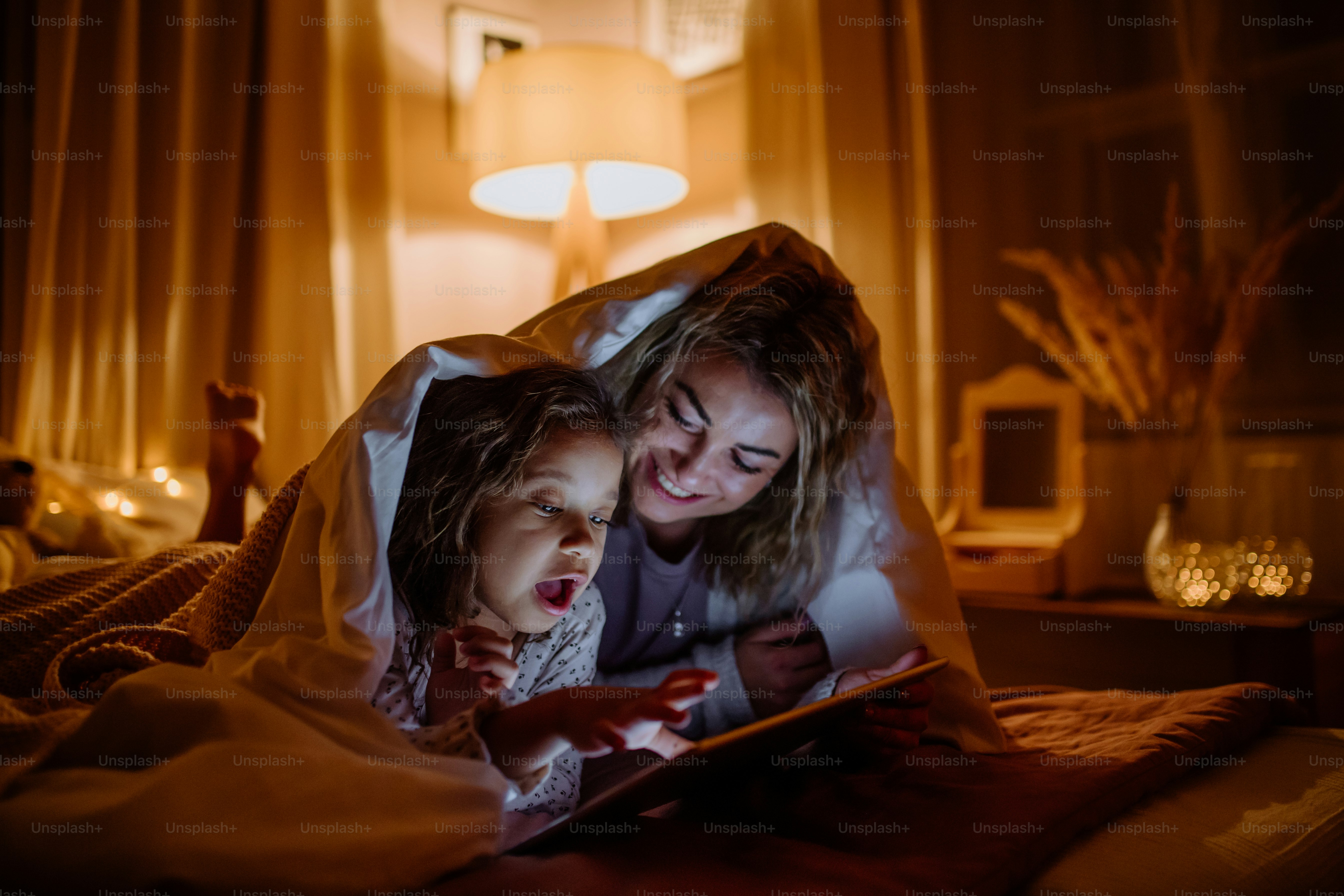 幼い娘が毛布の下に横たわり、家でタブレットで映画を見ている幸せな母親。