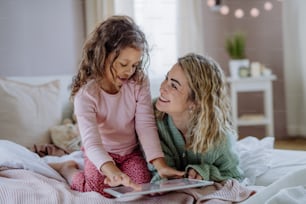 A happy mother with her little daughter sitting on bed and using tablet at home.