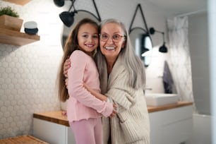 Eine ältere Großmutter und eine Enkelin stehen drinnen im Badezimmer, umarmen sich und schauen in die Kamera.