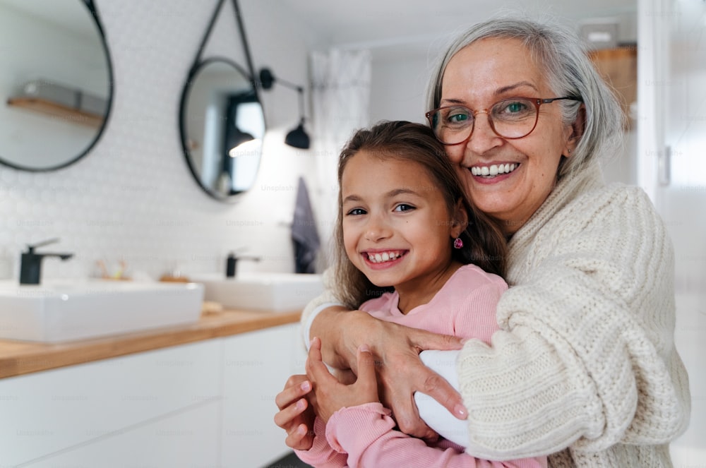 A senior grandmother and granddaughter standing indoors in bathroom, hugging and looking at camera.