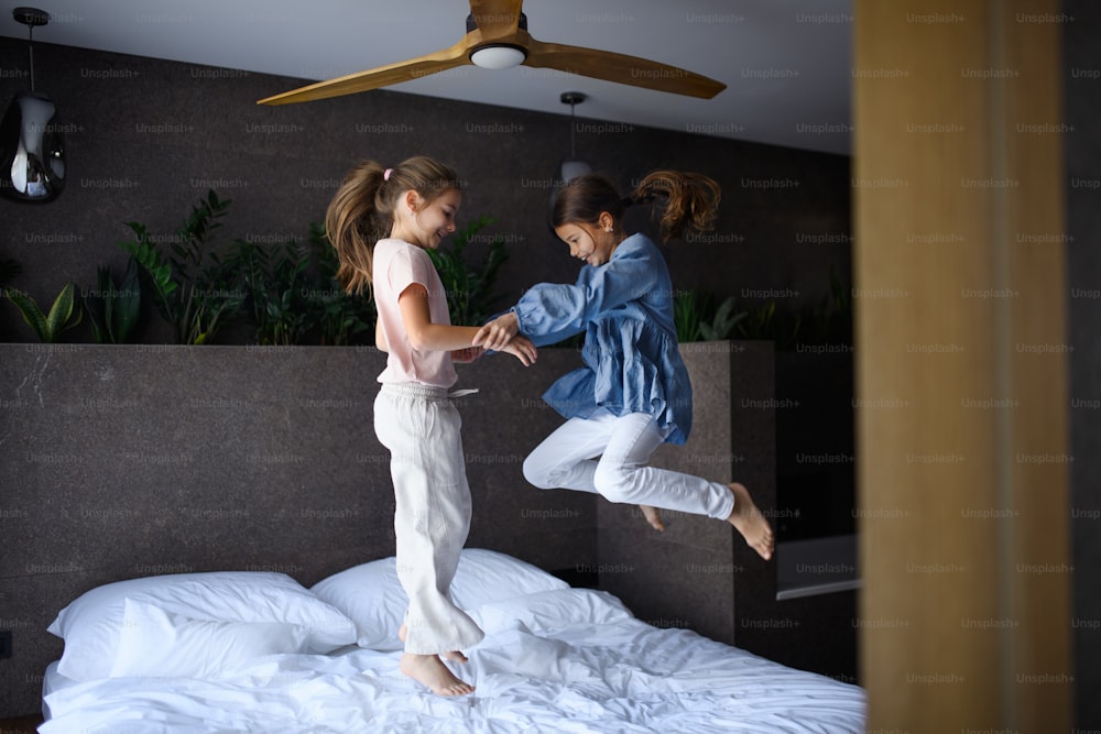 Two little sister holding hands and jumping on a bed indoors in hotel.