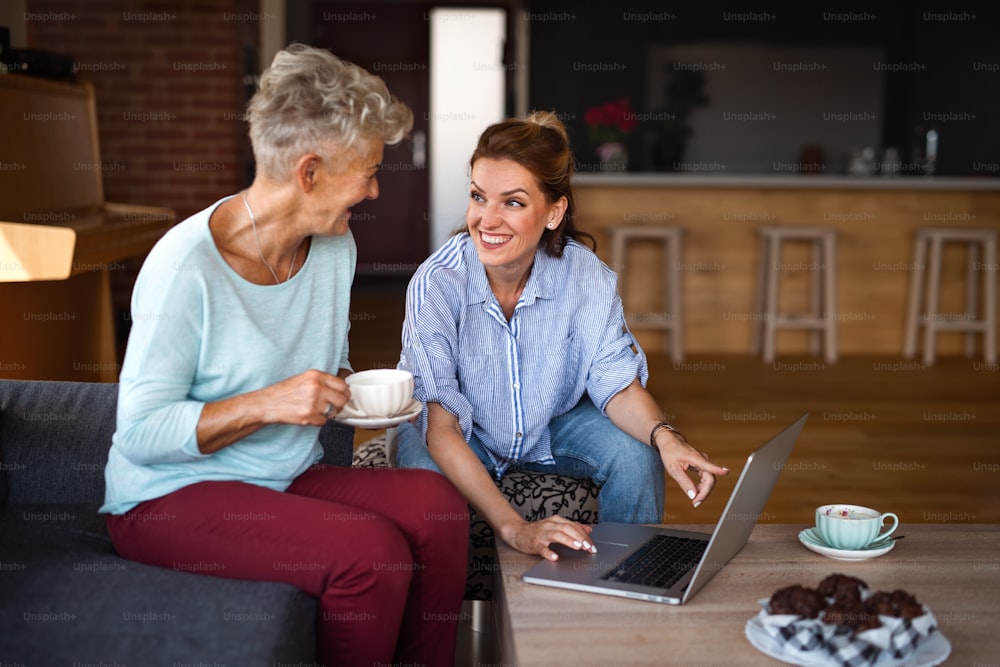 Une mère âgée heureuse prenant un café avec sa fille adulte à l’intérieur à la maison, assise, parlant et utilisant une tablette.