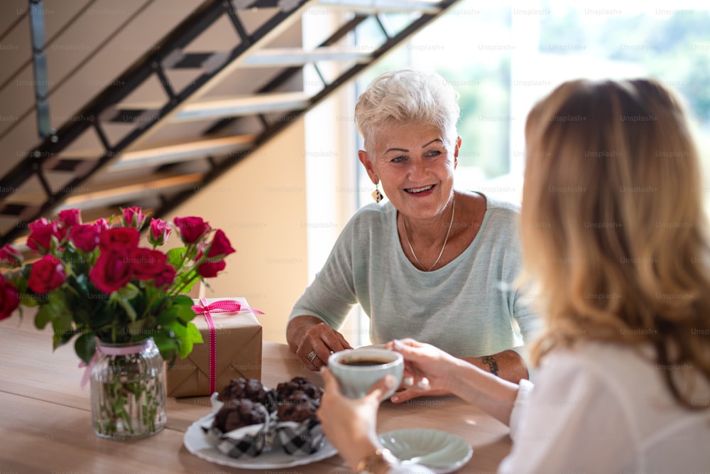 Une mère âgée heureuse prenant un café avec sa fille adulte à l’intérieur à la maison, assise et parlant.