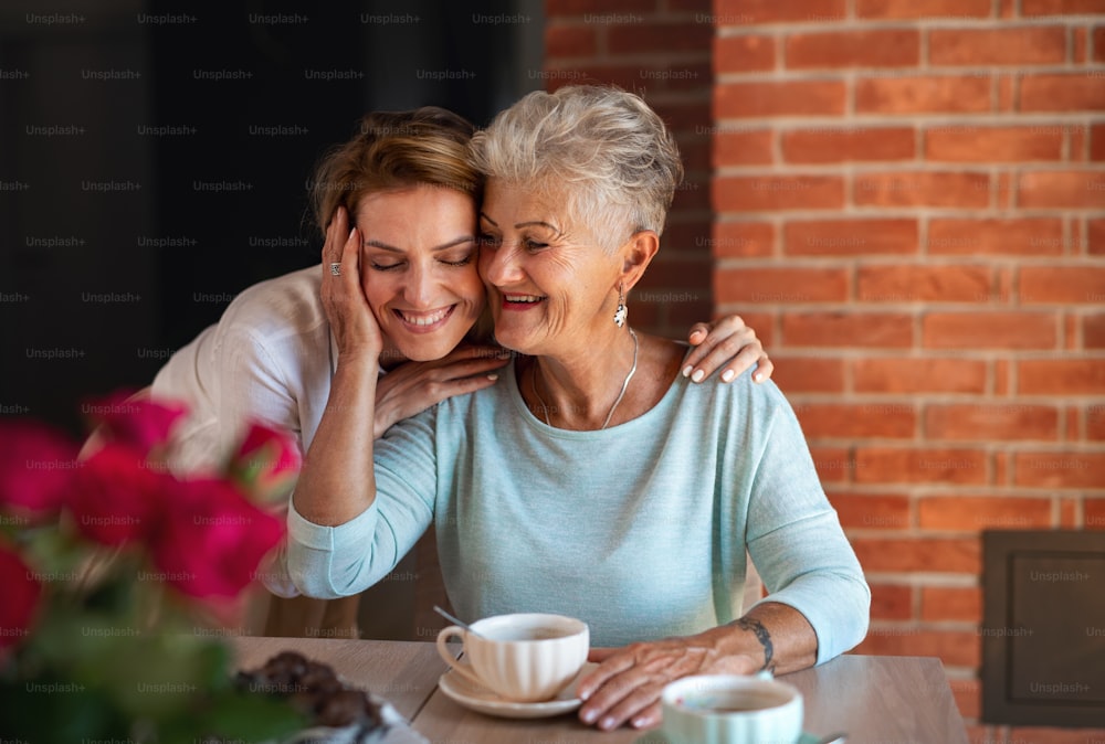 A happy senior mother embracing with adult daughter when sitting and having coffee indoors at home.