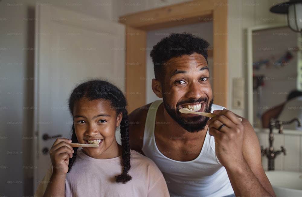 Un padre joven y feliz con una hija pequeña cepillándose los dientes en el interior de casa, un estilo de vida sostenible.