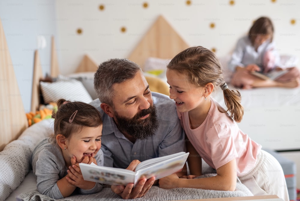A father with three daughters indoors at home, reading a book.