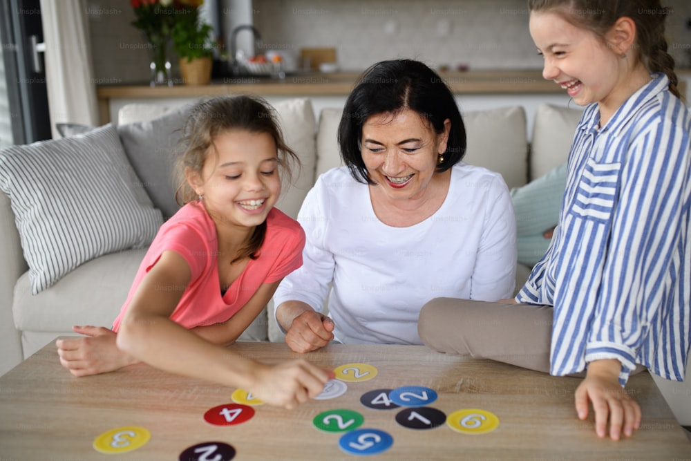 Niñas pequeñas felices con una abuela jugando a las cartas en el interior de casa.