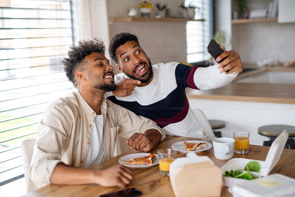 Un ritratto di giovani fratelli adulti in cucina al chiuso a casa, mentre scattano selfie.