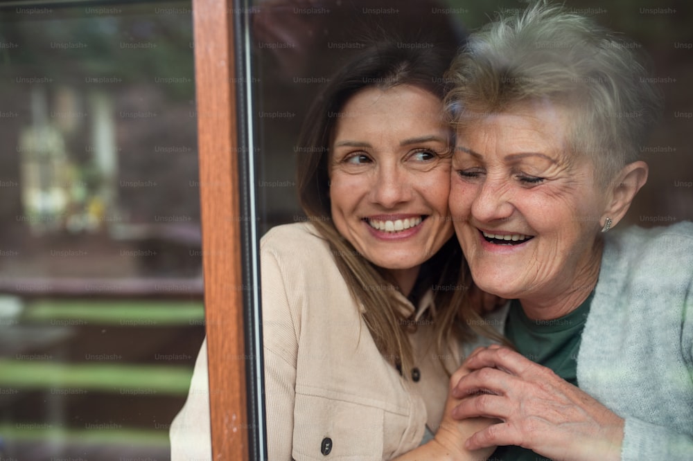A close-up portrait of happy senior mother with adult daughter indoors at home, hugging. Shot through window.