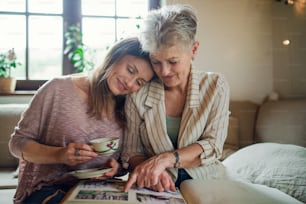 A senior mother with adult daughter indoors at home, looking at family photographs.