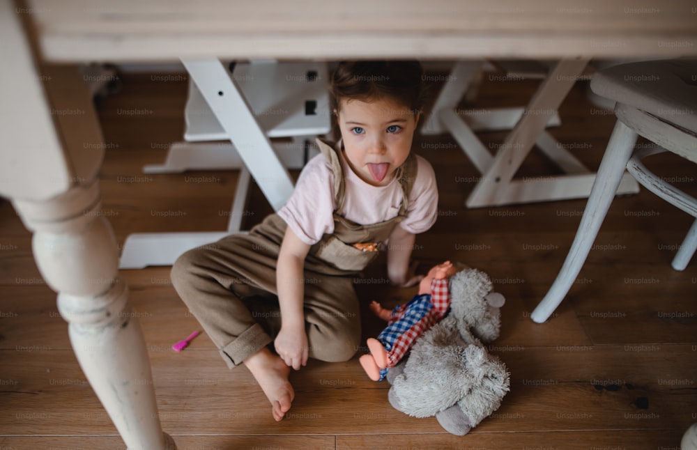 Un retrato de una linda niña sentada en el suelo debajo de la mesa en el interior de su casa, sacando la lengua.