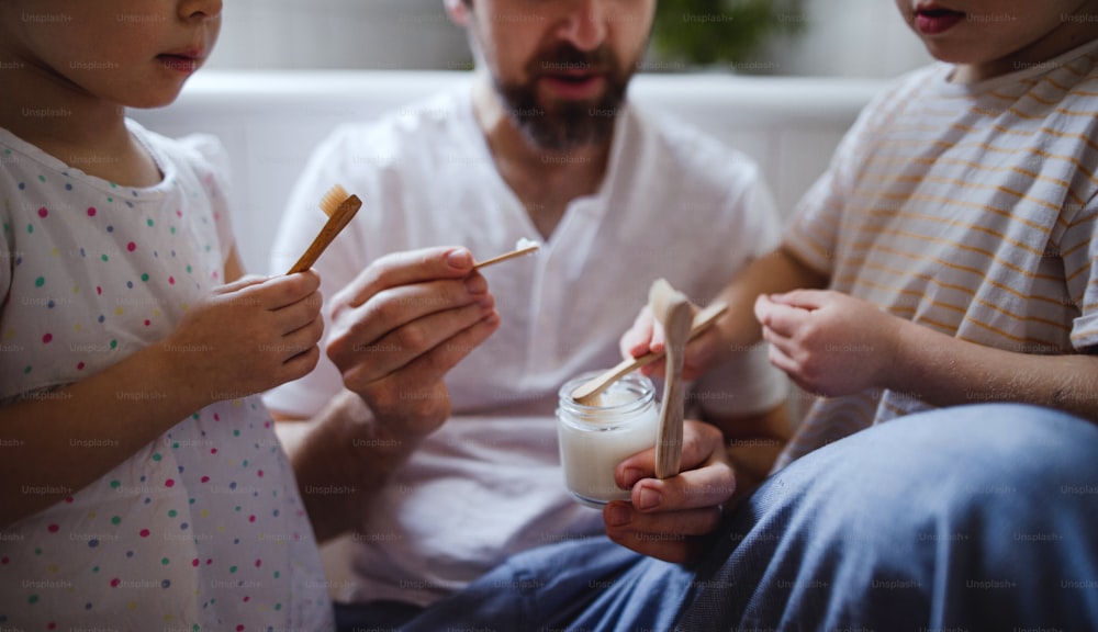 Um pai irreconhecível com dois filhos pequenos escovando os dentes dentro de casa, conceito de estilo de vida sustentável.