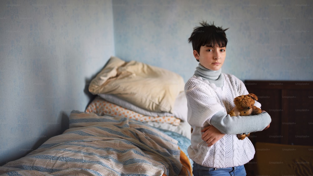 Poor sad small girl with teddy bear standing and looking at camera indoors at home, poverty concept.