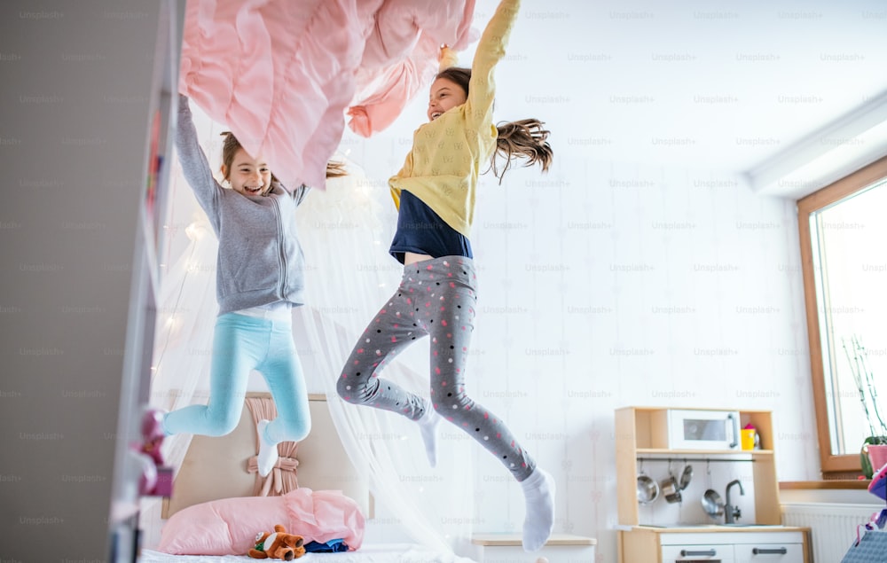Dos alegres hermanas niñas pequeñas en el interior de casa, saltando sobre la cama en el dormitorio.