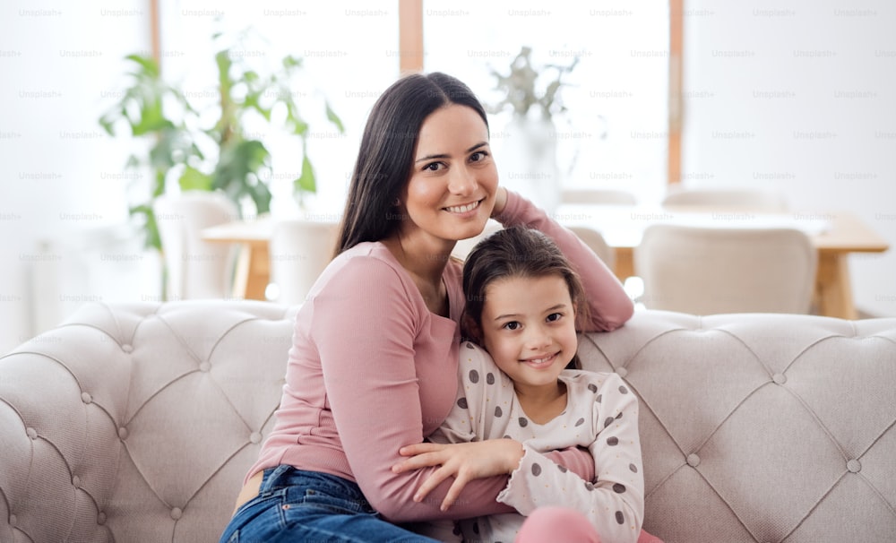 Portrait of mother with small daughter sitting on sofa indoors at home, looking at camera.