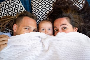 Portrait of young couple with baby daughter resting under blanket, looking at camera.
