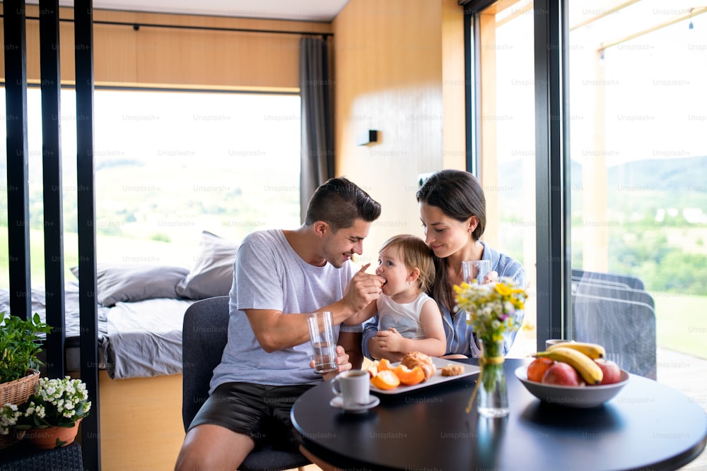 Young couple with small daughter eating breakfast indoors, weekend away in container house in countryside.