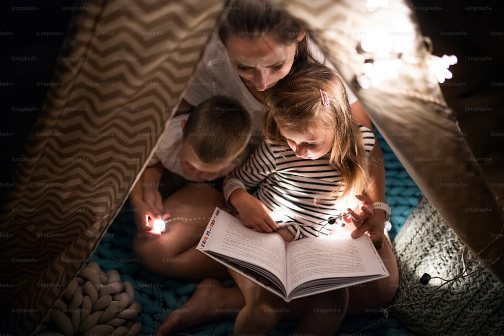 Draufsicht auf Mutter mit Kindern, die drinnen im Schlafzimmer sitzt und ein Buch liest.