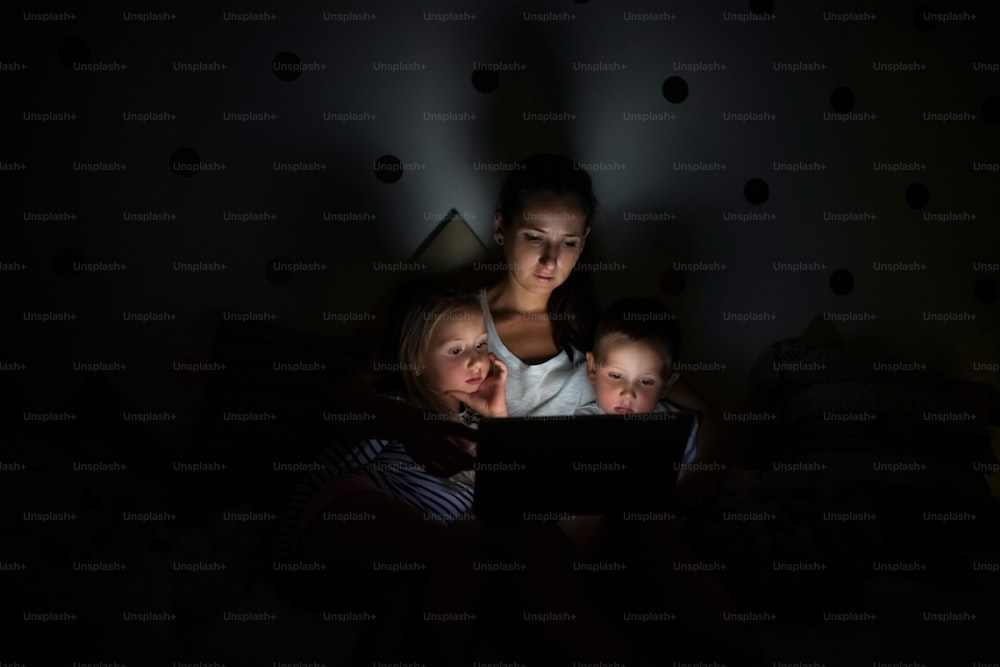 Junge Mutter mit Kindern, die abends im Schlafzimmer sitzen und einen Laptop benutzen.