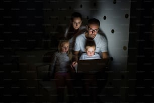 Front view of family with children sitting indoors in bedroom, using laptop.