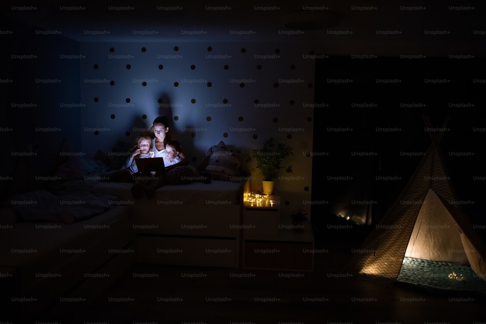 Mãe pequena com filhos sentados dentro de casa no quarto à noite, usando laptop.