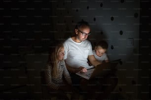 Father with small children sitting indoors in bedroom in the evening, using laptop.