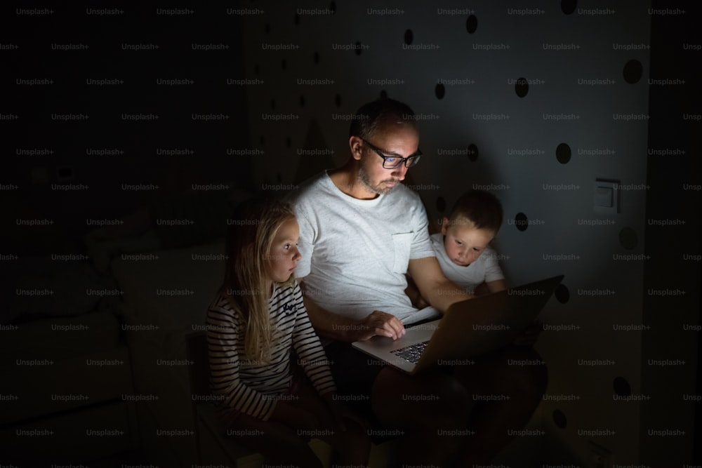 Père avec de jeunes enfants assis à l’intérieur dans la chambre le soir, en utilisant un ordinateur portable.