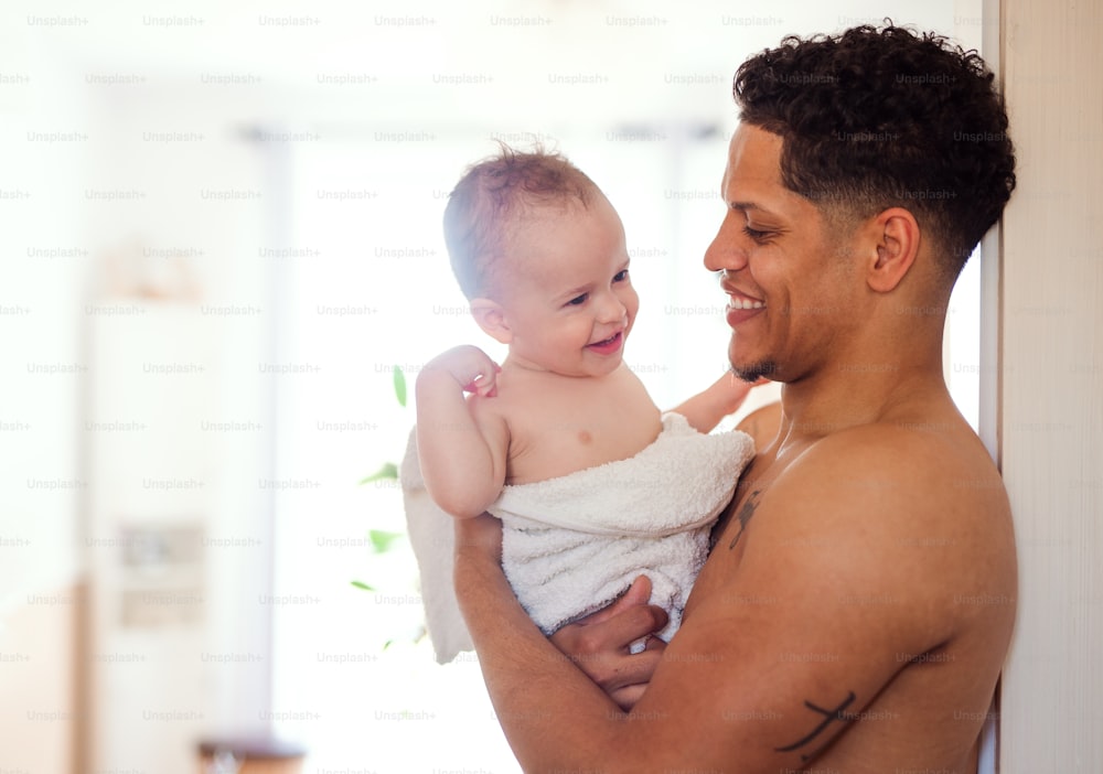Pai de topless e filho pequeno embrulhados em toalha em um banheiro dentro de casa.
