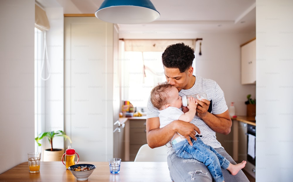 A cheerful father bottle feeding a small toddler son indoors at home.
