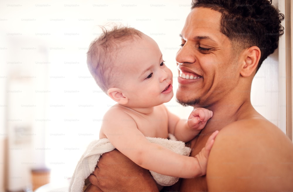 Topless father and small toddler son wrapped in towel in a bathroom indoors at home.