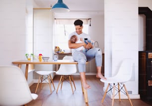 A cheerful father bottle feeding a small toddler son indoors at home.