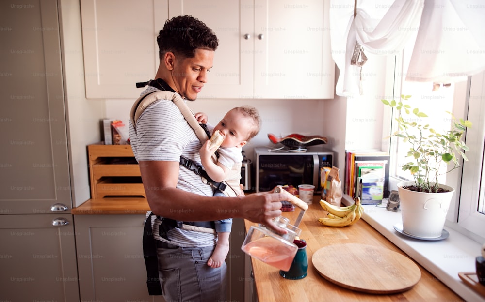 A father with small toddler son in carrier in kitchen indoors at home, pouring tea.
