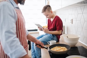 A mature father and small son with tablet indoors in kitchen, making pancakes.