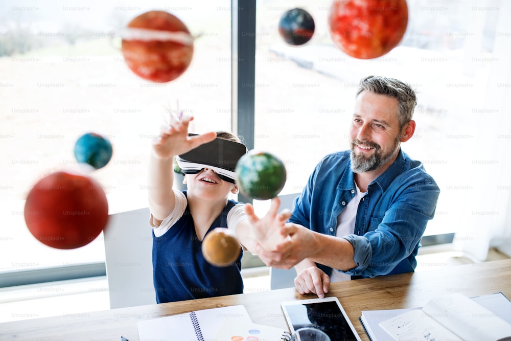 Ein reifer Vater mit kleinem Sohn und VR-Brille, der drinnen am Tisch sitzt und an einem Schulprojekt arbeitet.