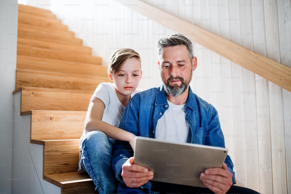 A mature father with small son sitting on the stairs indoors, using tablet.